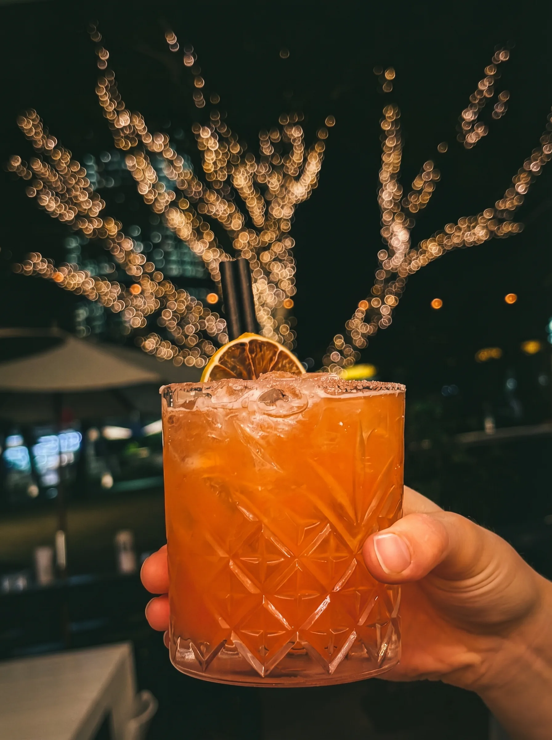 Aperol Margarita at Poppies Southbank — amber crystal glass with dried lime garnish, South Bank fairy lights in background